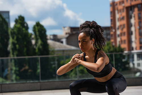 African american sportswoman in earphone doing squat while training outdoors