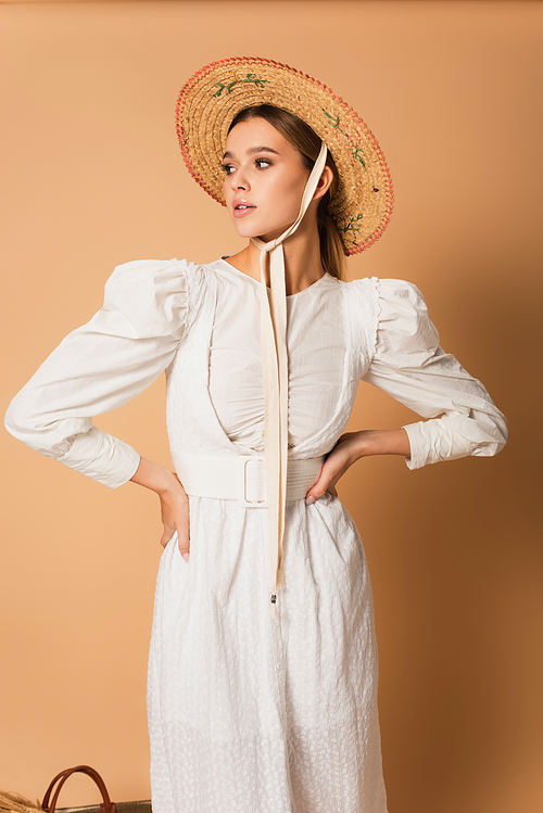 young woman in white dress and straw hat standing with hands on hips on beige