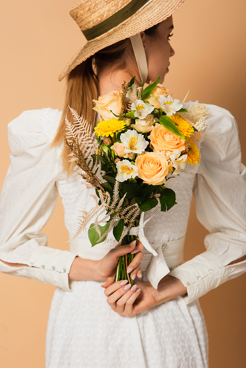 young woman in straw hat holding bouquet of flowers behind back on beige