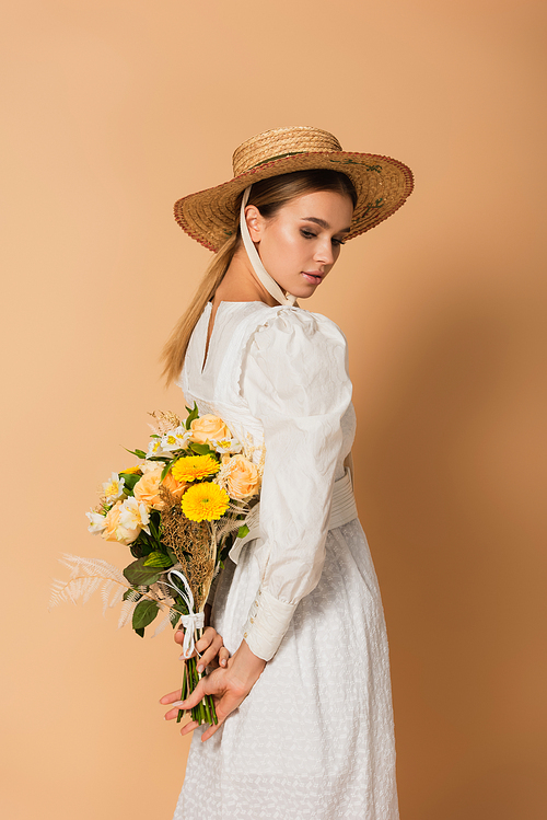 young woman in dress and straw hat holding bouquet of flowers behind back on beige