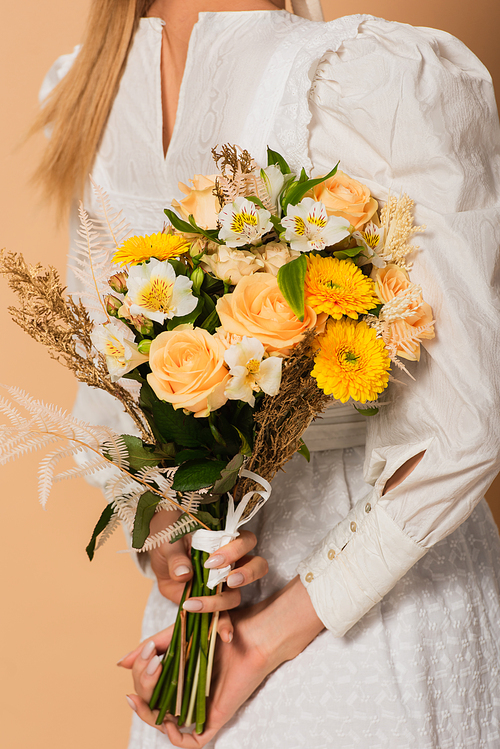 cropped view of young woman in dress holding bouquet of flowers behind back on beige