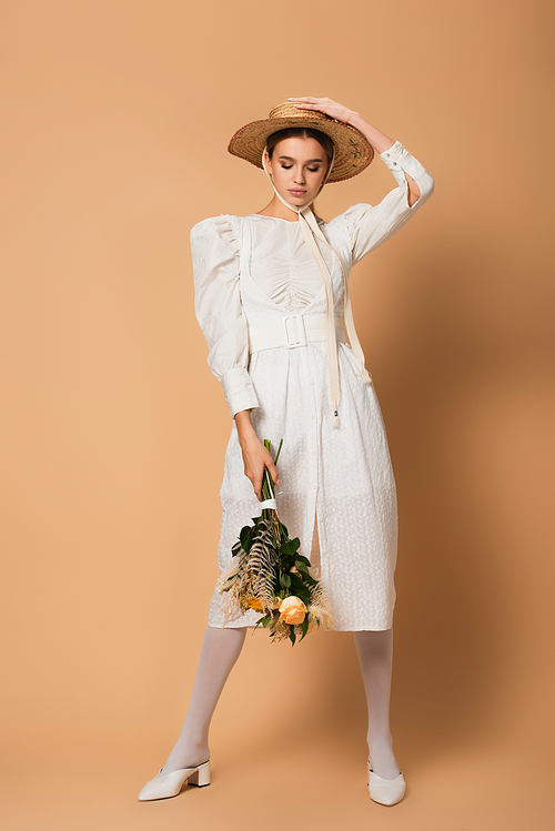 full length of young woman in dress and straw hat holding bouquet of flowers on beige