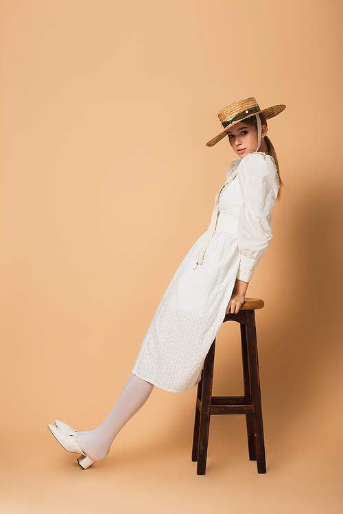 full length of young woman in white dress and straw hat leaning on stool on beige