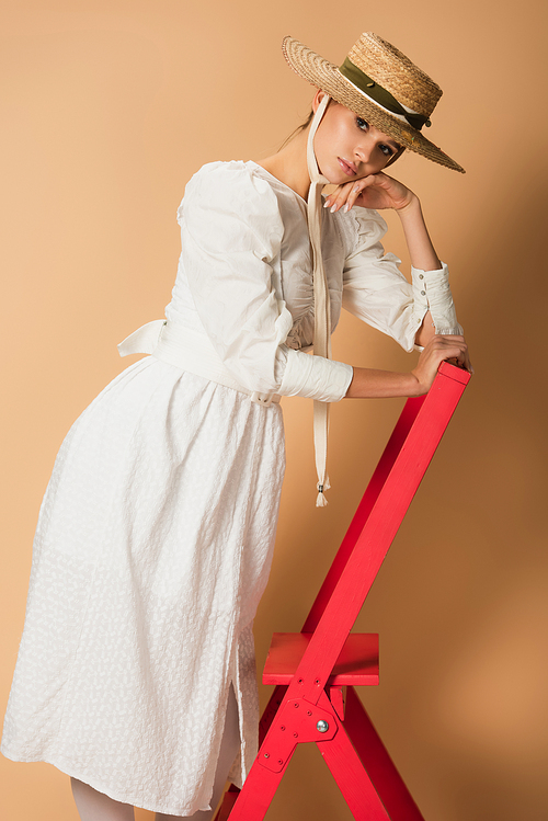 young woman in white dress and straw hat standing on red ladder on beige