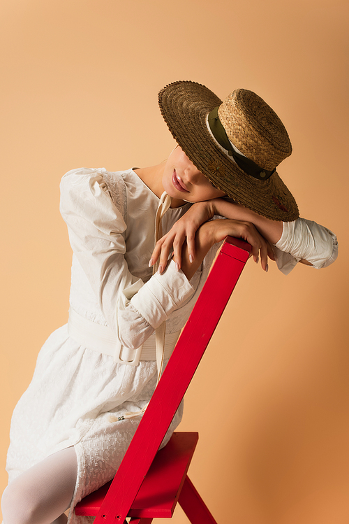 young woman in white dress and straw hat leaning on red ladder on beige