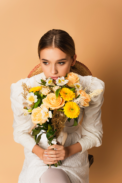 young woman in white dress holding bouquet with different flowers on beige