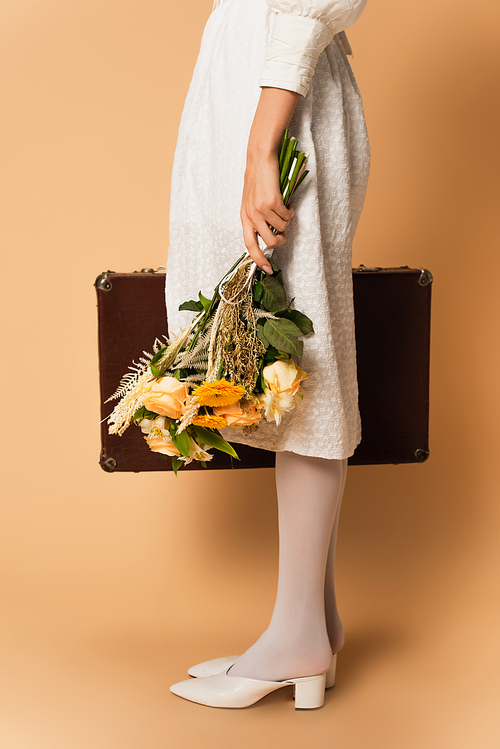 cropped view of young woman in dress holding bouquet of flowers and suitcase on beige