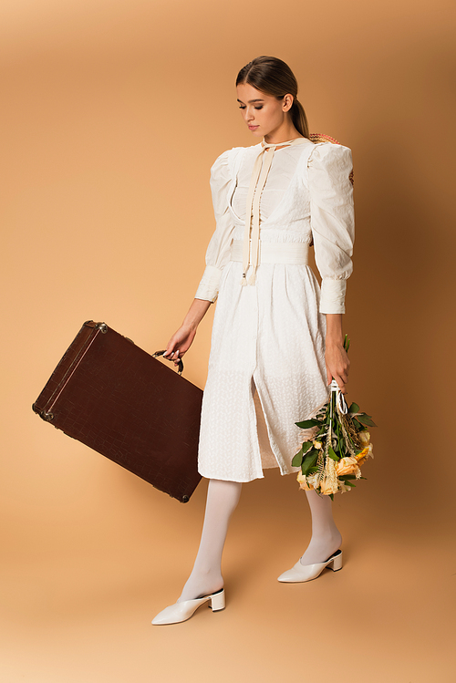 full length of young woman in white dress holding bouquet with flowers and suitcase on beige