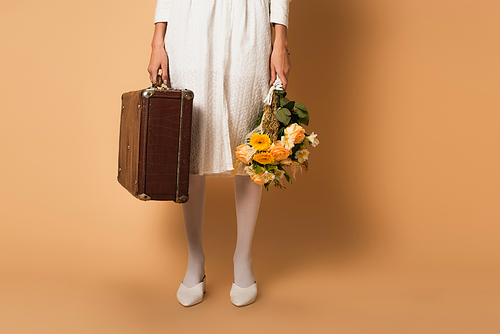 cropped view of young woman in white dress holding bouquet of flowers and suitcase on beige