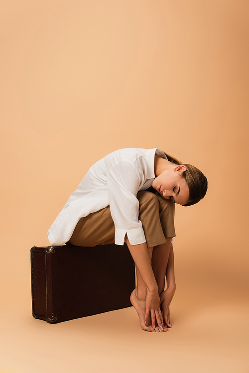 barefoot woman in white shirt and pants sitting on vintage suitcase with closed eyes on beige background