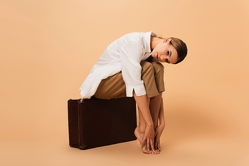 barefoot woman in shirt and trousers touching legs while sitting on retro suitcase on beige background