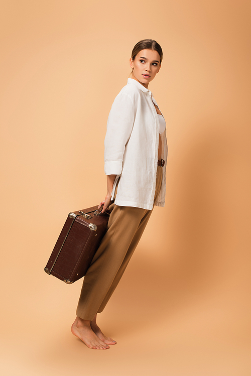 barefoot woman in pants and white shirt standing on tiptoe with vintage suitcase on beige background