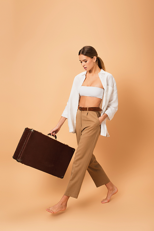 young woman in pants and unbuttoned shirt walking barefoot with vintage suitcase on beige background