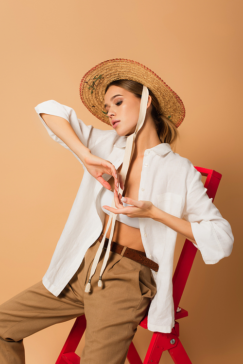 sensual woman in unbuttoned shirt and straw hat posing on ladder on beige background