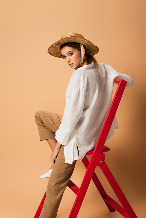trendy woman in white shirt, pants and straw hat  on red ladder on beige background