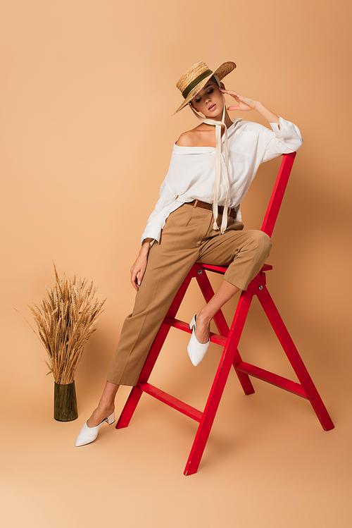 pretty woman in white shirt, pants and straw hat posing on red ladder near vase with spikelets on beige