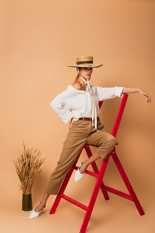 pretty woman in white shirt, pants and straw hat posing on ladder with hand on hip on beige background