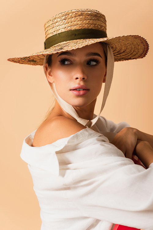 pretty woman in white shirt and straw hat posing with naked shoulder isolated on beige