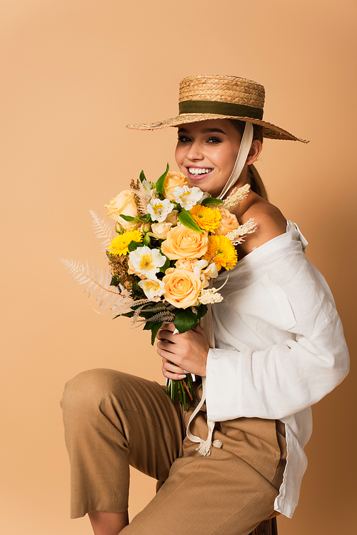 smiling young woman in white shirt and straw hat holding bouquet of flowers on beige