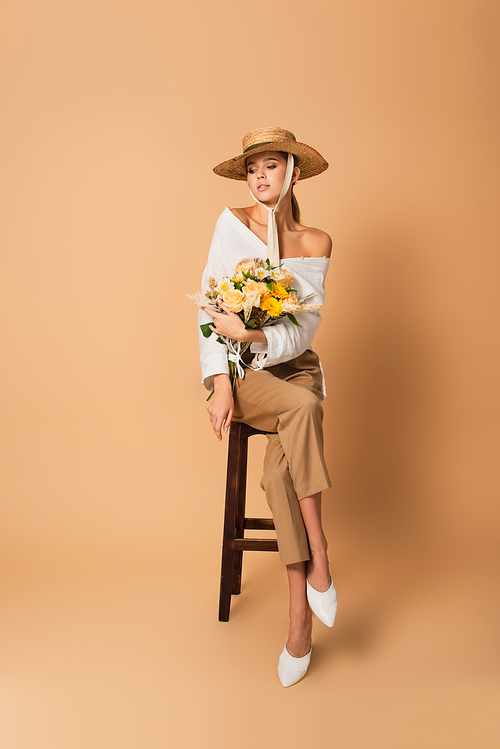 full length of young woman in straw hat holding bouquet of different flowers and sitting on wooden stool on beige