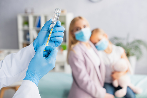 Pediatrician in latex gloves picking up vaccine near blurred family in hospital