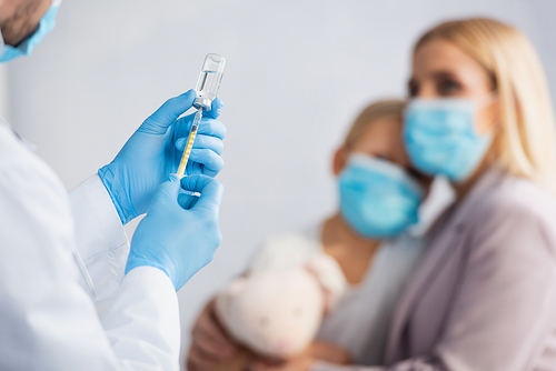 Doctor in latex gloves picking up vaccine in syringe near mother and kid on blurred background