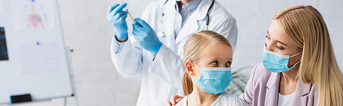 Mother and kid in medical masks near blurred family doctor with vaccine, banner