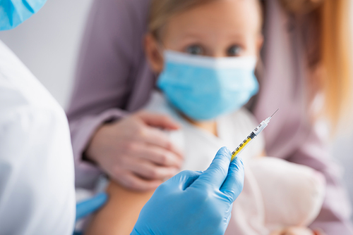 Doctor holding syringe near kid and woman on blurred background