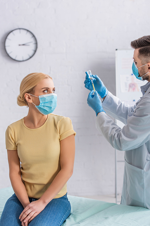 woman in protective mask looking at doctor filling syringe with vaccine