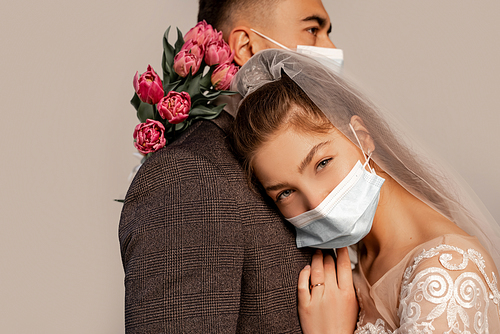 young bride in medical mask leaning on chest of groom and  isolated on grey