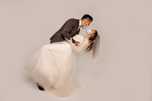 side view of newlyweds in medical masks dancing isolated on grey with lilac shade