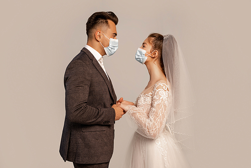 side view of groom and bride in medical masks holding hands isolated on grey with lilac shade