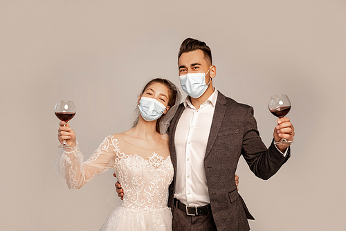young newlyweds in medical masks holding glasses with red wine isolated on grey