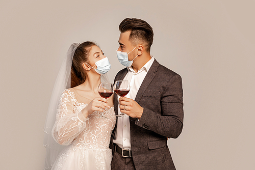newlyweds in safety masks looking at each other while clinking wine glasses isolated on grey