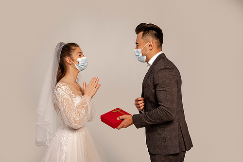 side view of man in medical mask presenting gift box to elegant bride isolated on grey