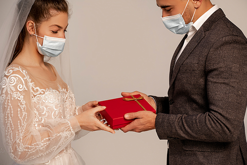 man in medical mask presenting gift box to young bride isolated on grey
