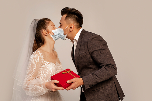 groom in medical mask kissing bride while presenting gift isolated on grey