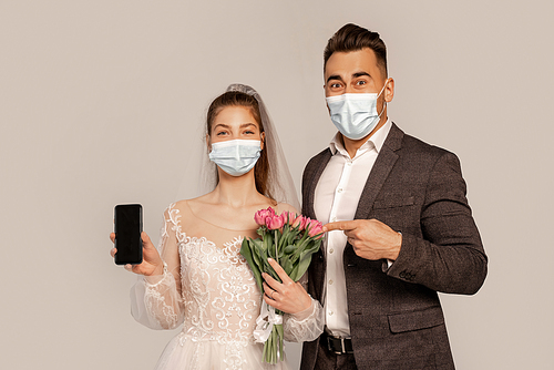 man in medical mask pointing at smartphone in hand of bride isolated on grey