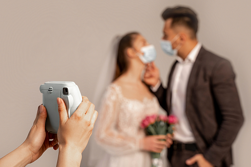woman taking photo of blurred newlyweds in medical masks isolated on grey