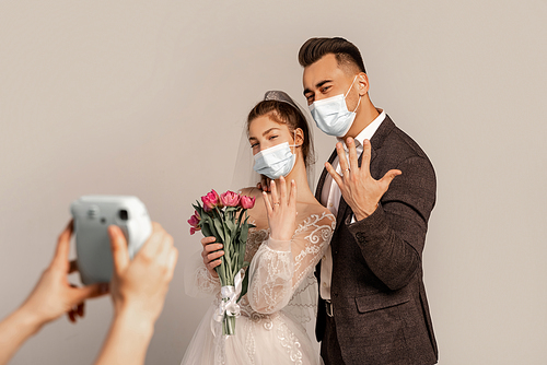 newlyweds in medical masks showing wedding rings near blurred photographer isolated on grey
