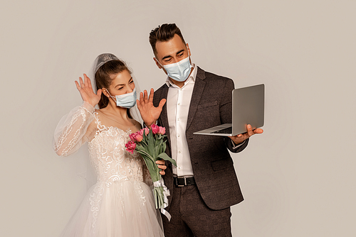 bride and groom in medical masks waving hands during video call isolated on grey