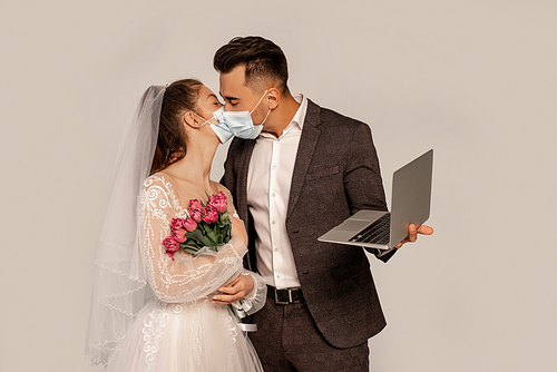 man in medical mask kissing bride while holding laptop isolated on grey
