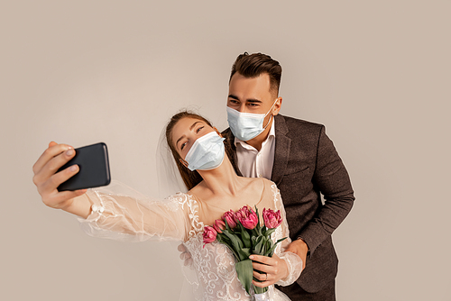newlyweds in medical masks taking selfie on mobile phone isolated on grey