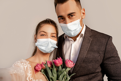 young newlyweds in medical masks  near fresh tulips isolated on grey