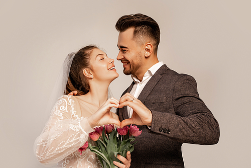 joyful newlyweds smiling at each other while showing heart symbol with hands isolated on grey