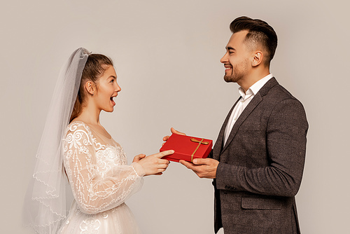 side view of happy man presenting gift to amazed bride isolated on grey
