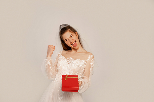 excited bride showing yeah gesture while holding present isolated on grey