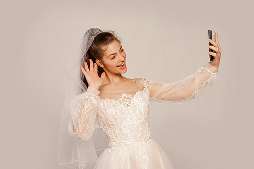 happy bride waving hand while taking selfie isolated on grey with lilac shade