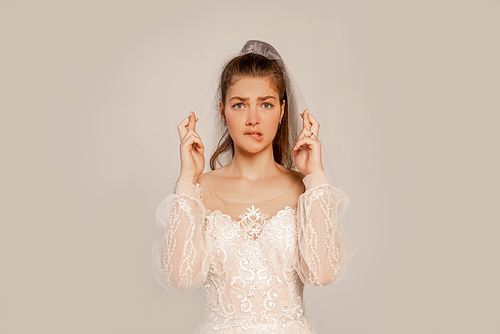 worried woman in wedding dress with crossed fingers isolated on grey