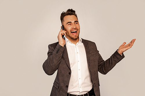 excited man gesturing while talking on smartphone isolated on grey